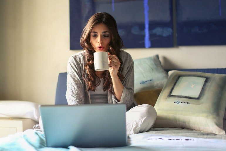 Woman on bed with laptop sipping coffee