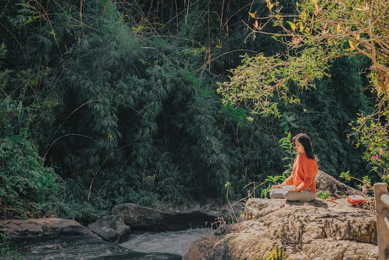 A woman sitting on a rock by a stream meditating