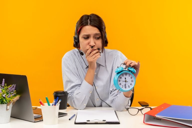 A woman sat at a cluttered desk holding a blue alarm clock