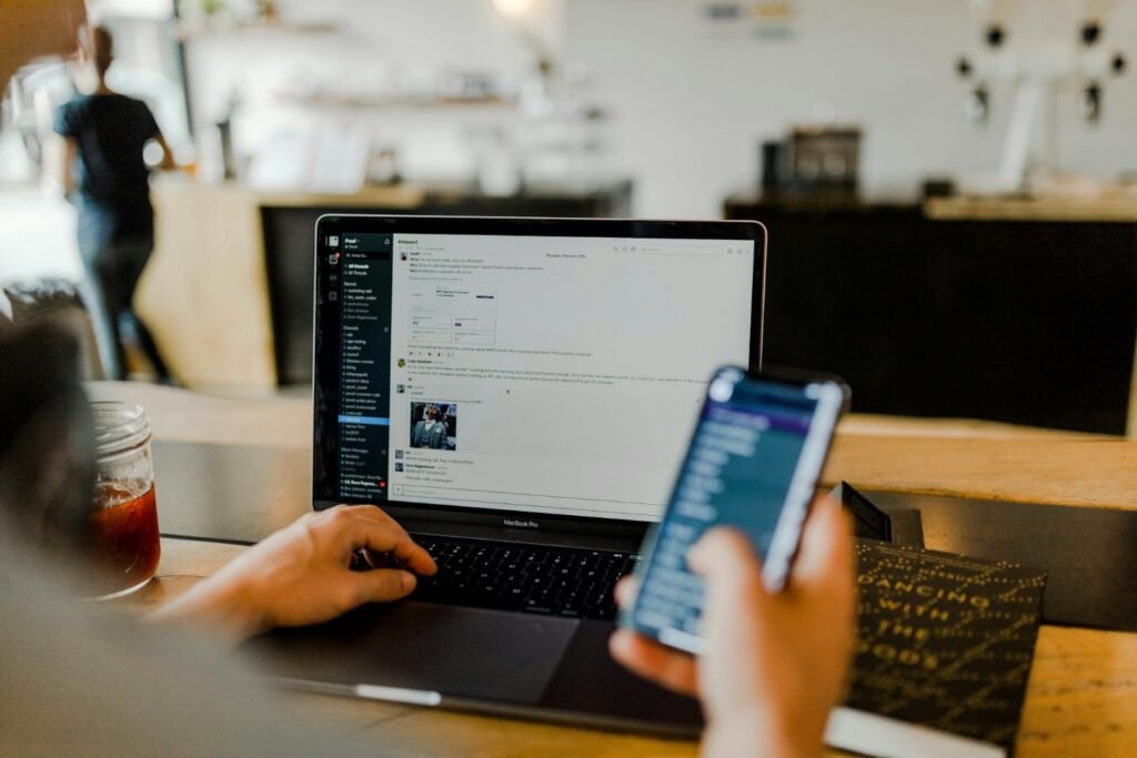 A person holding a mobile phone while looking at a laptop screen