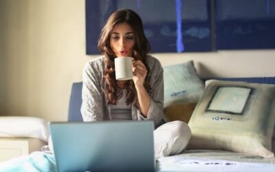 A woman sitting on a bed, drinking a cup of coffee and viewing a laptop